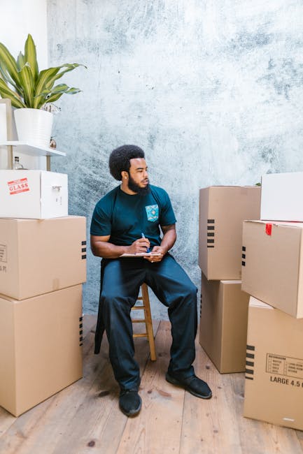 A young man with curly dark hair, glasses, and a beard sits inside a large indoor space, likely a storage or packing area, surrounded by stacked cardboard boxes and large wrapped furniture items. He i